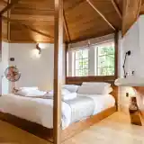 A cozy bedroom featuring a large wooden bed with white bedding, complemented by a light gray throw. Wooden beams and a warm color palette enhance the inviting atmosphere. Natural light streams in through large windows, with modern light fixtures and a decorative fan in the corner.
