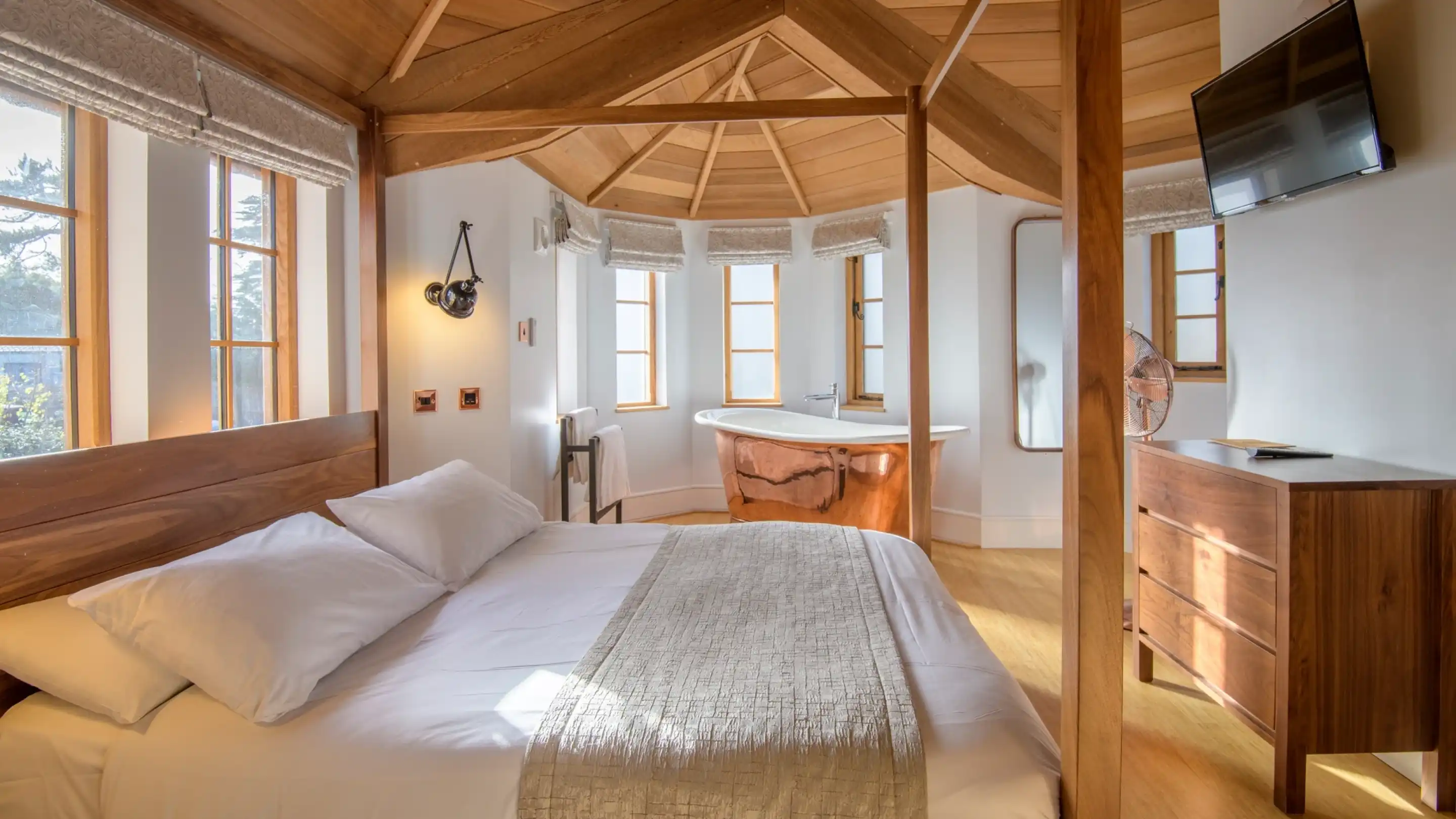 A cozy bedroom featuring a wooden ceiling with exposed beams, a large bed with white linens, and modern decor. A stylish curved bathtub is visible beside a window, complemented by warm sunlight filtering through. A dresser and a wall-mounted television complete the space.