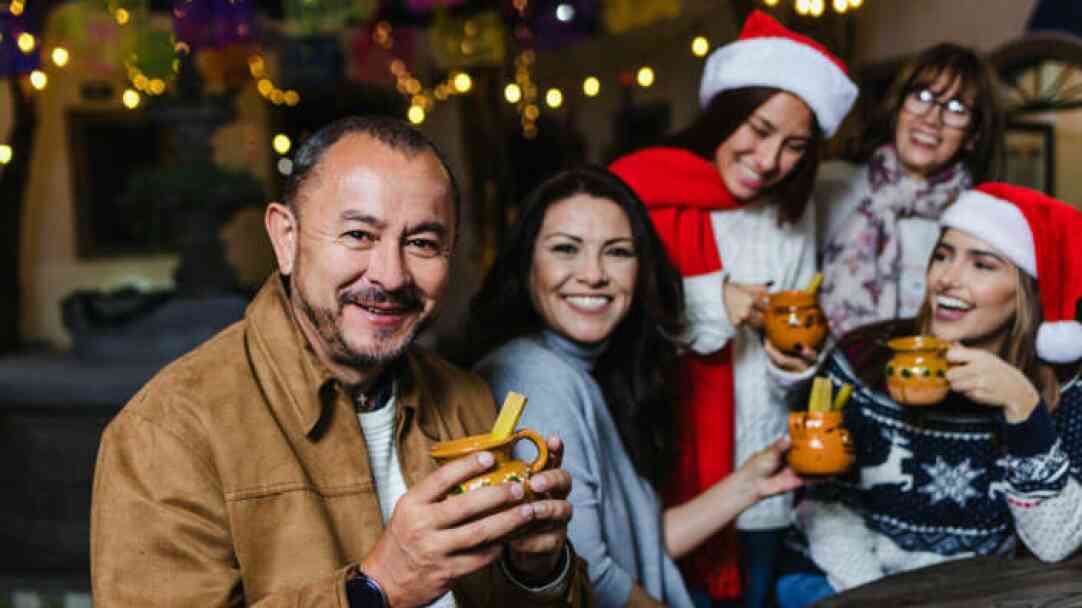 A group of five people, wearing festive attire including Santa hats, smiles while holding drinks. They are in a decorated setting with colorful lights and festive decorations.