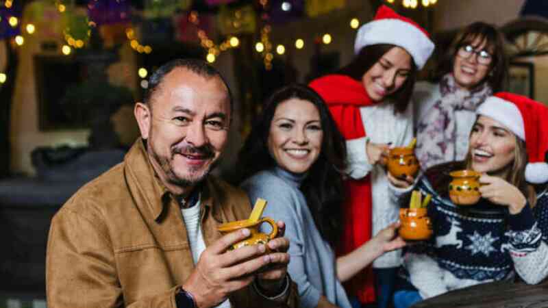 A group of five people, wearing festive attire including Santa hats, smiles while holding drinks. They are in a decorated setting with colorful lights and festive decorations.
