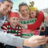 A family smiles while taking a selfie together. They are wearing festive holiday sweaters and paper crowns. The atmosphere is cheerful and cozy, with a window and soft lighting in the background.
