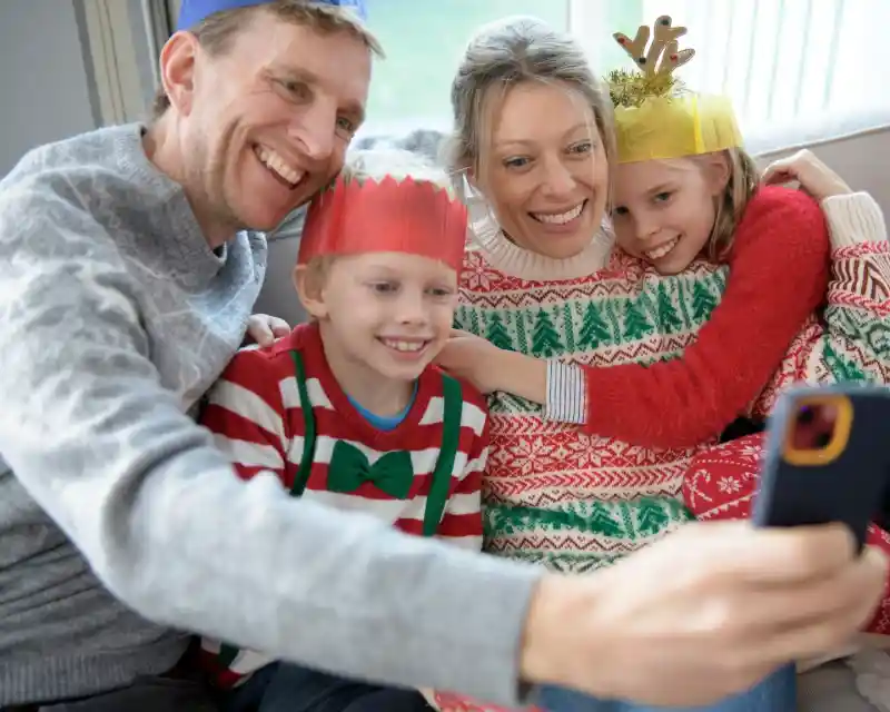 A family smiles while taking a selfie together. They are wearing festive holiday sweaters and paper crowns. The atmosphere is cheerful and cozy, with a window and soft lighting in the background.