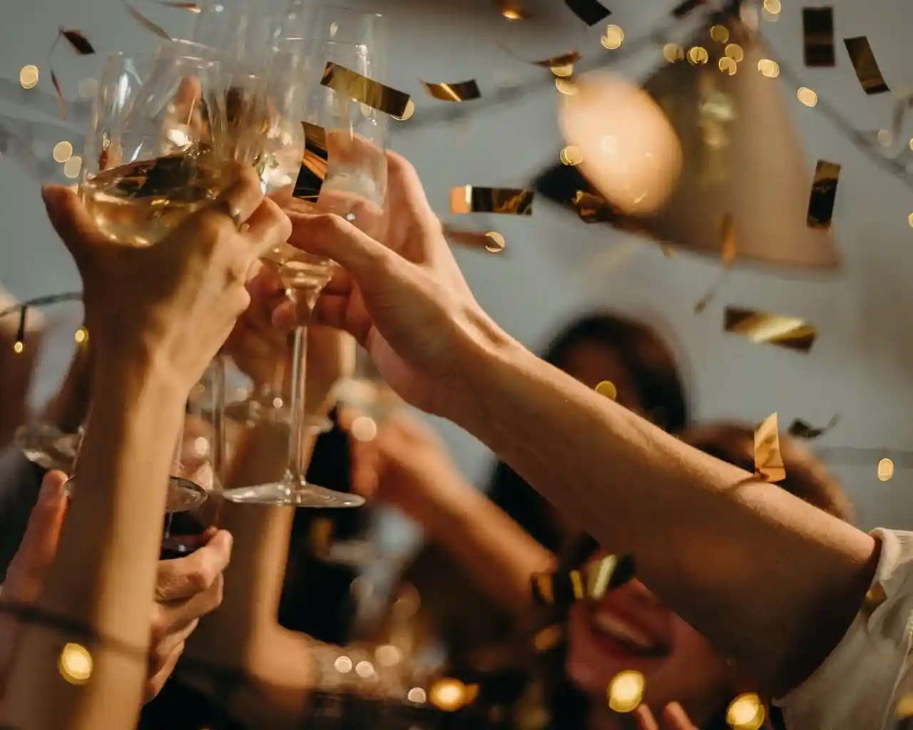 Glasses of champagne clinking together in celebration, surrounded by golden confetti and festive lights. Happy faces are partially visible in the background, adding to the joyful atmosphere.