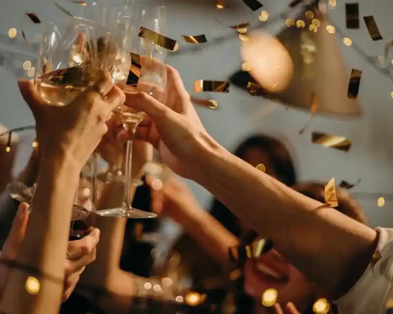 Glasses of champagne clinking together in celebration, surrounded by golden confetti and festive lights. Happy faces are partially visible in the background, adding to the joyful atmosphere.