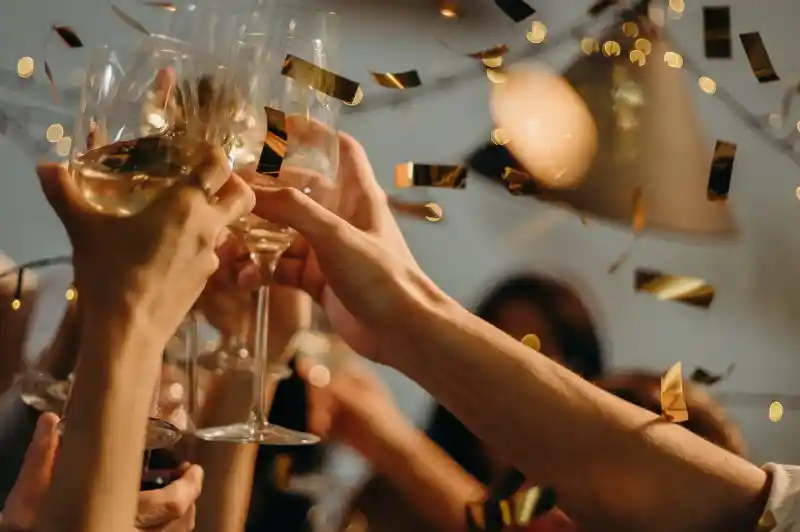 Glasses of champagne clinking together in celebration, surrounded by golden confetti and festive lights. Happy faces are partially visible in the background, adding to the joyful atmosphere.