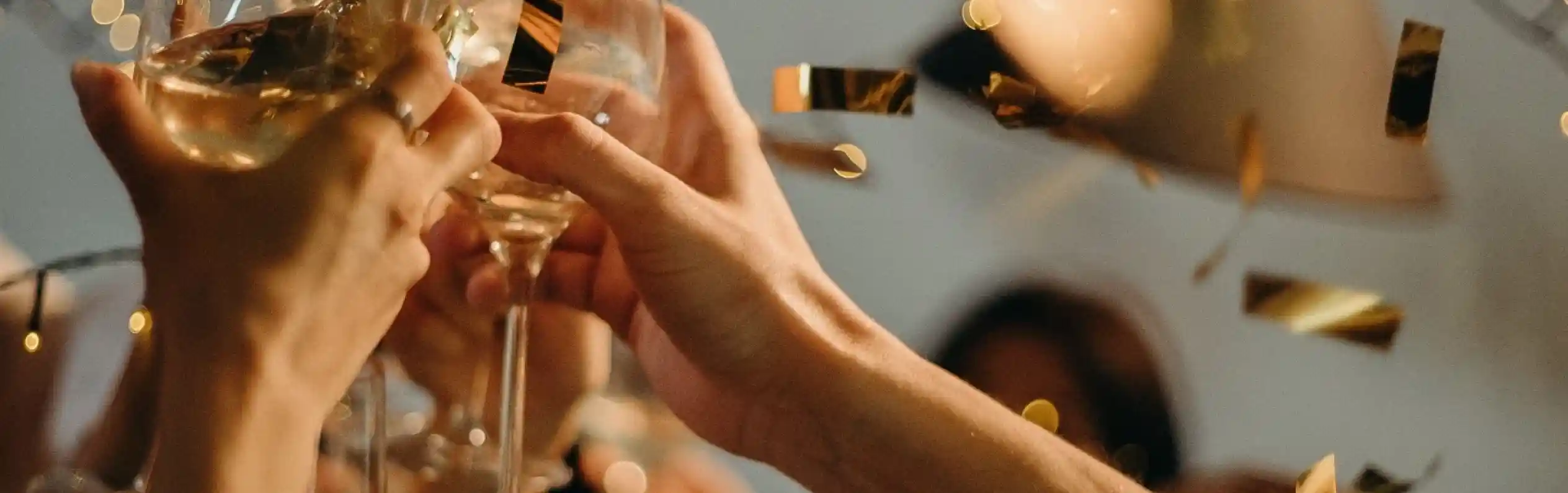 Glasses of champagne clinking together in celebration, surrounded by golden confetti and festive lights. Happy faces are partially visible in the background, adding to the joyful atmosphere.