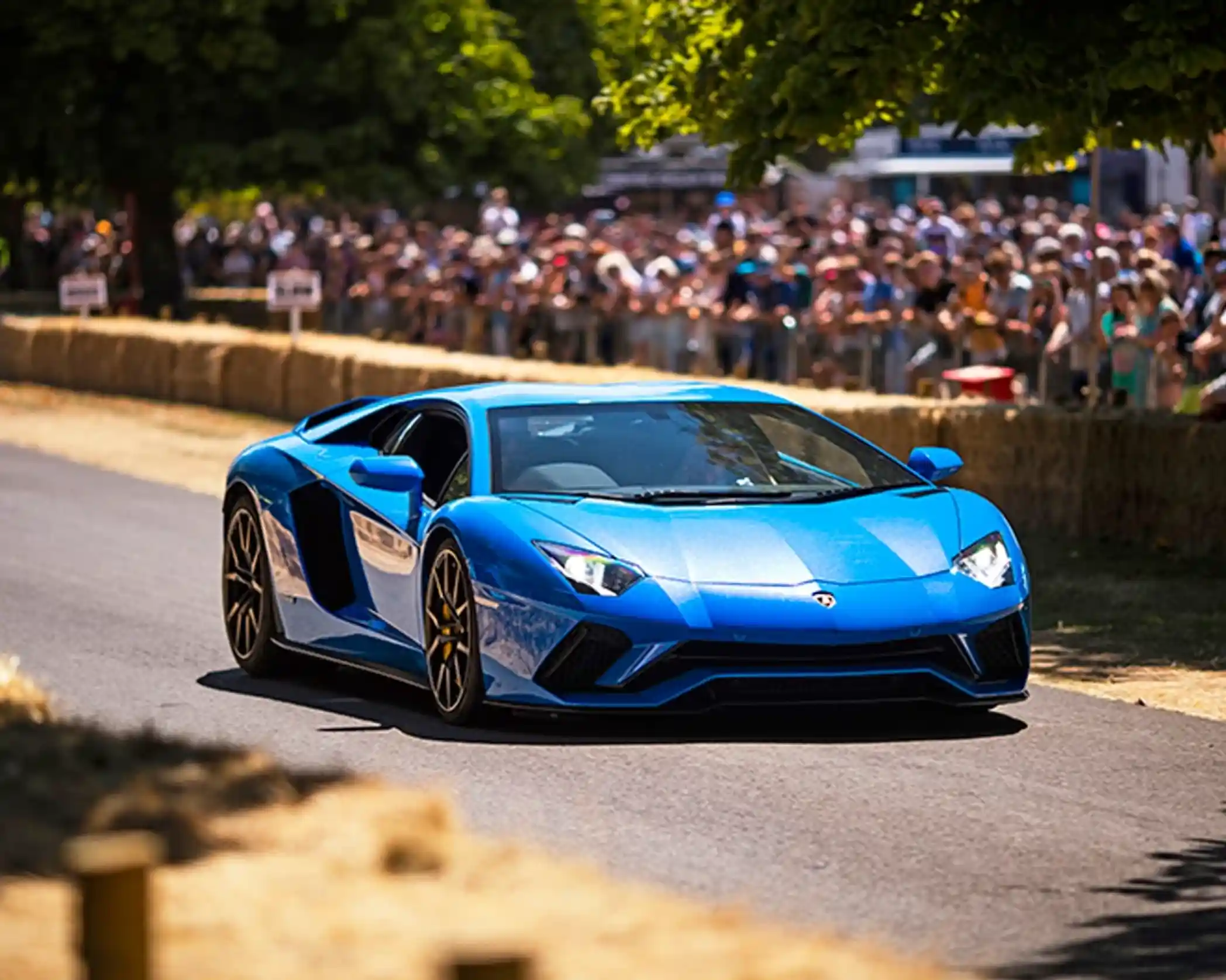 A vibrant blue Lamborghini Aventador cruising down a country road, with a crowd of spectators watching from the sidelines. Sunlight filters through the trees, highlighting the car's sleek design.