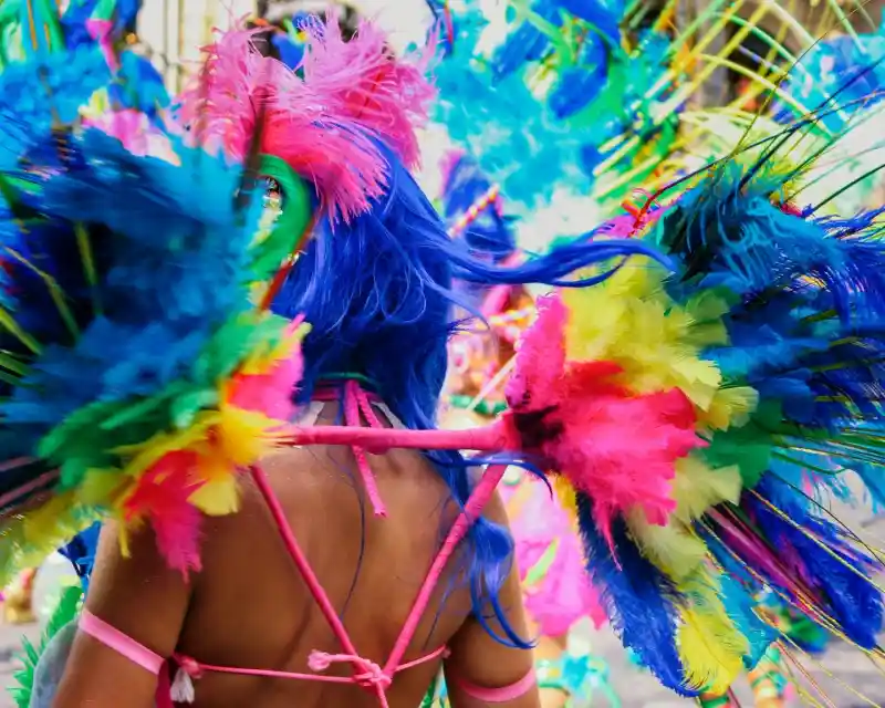 A person with vibrant blue hair is seen from behind, adorned with colorful feathers in bright shades of pink, blue, and yellow. The elaborate costume features feathered wings, creating a lively and festive atmosphere.