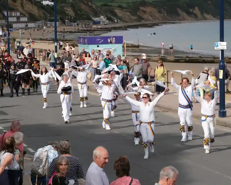 A vibrant parade scene along a seaside promenade. A group of performers in white outfits with colorful accents marches joyfully, waving their arms. Crowds of spectators line the street, watching the event against a backdrop of the beach and cliffs.