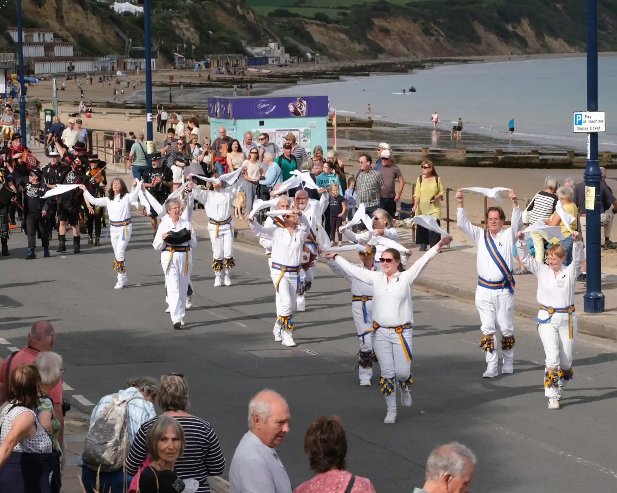 A vibrant parade scene along a seaside promenade. A group of performers in white outfits with colorful accents marches joyfully, waving their arms. Crowds of spectators line the street, watching the event against a backdrop of the beach and cliffs.