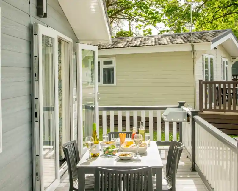 A cozy outdoor dining area on a deck, featuring a table set with wine, glasses, and a spread of food, surrounded by greenery and nearby cabins.