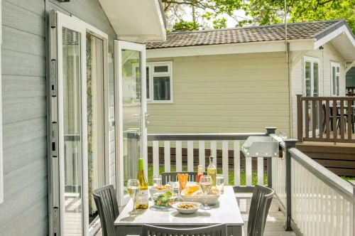 A cozy outdoor dining area on a deck, featuring a table set with wine, glasses, and a spread of food, surrounded by greenery and nearby cabins.