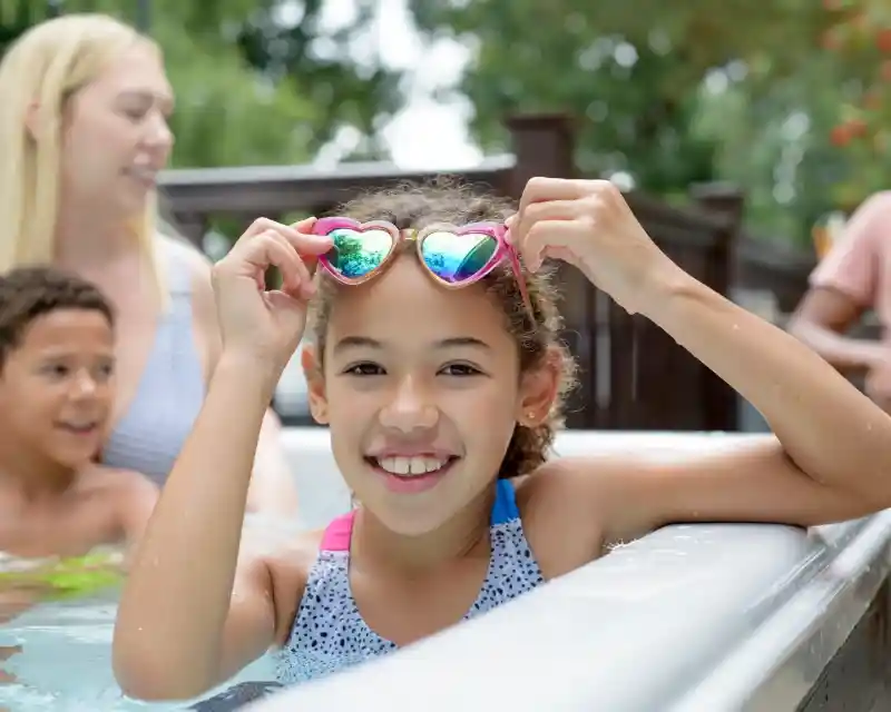 A young girl with curly hair smiles while holding heart-shaped sunglasses at the edge of a pool. In the background, a woman and a boy are partially visible, enjoying the water. Bright greenery surrounds the scene.
