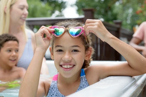 A young girl with curly hair smiles while holding heart-shaped sunglasses at the edge of a pool. In the background, a woman and a boy are partially visible, enjoying the water. Bright greenery surrounds the scene.