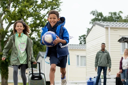 Two children are joyfully running down a path; a boy carries a soccer ball while wearing a blue jacket and shorts. A girl follows, pulling a suitcase and wearing a green shirt with a smiley face. In the background, two adults stand by a parked car near mobile homes.