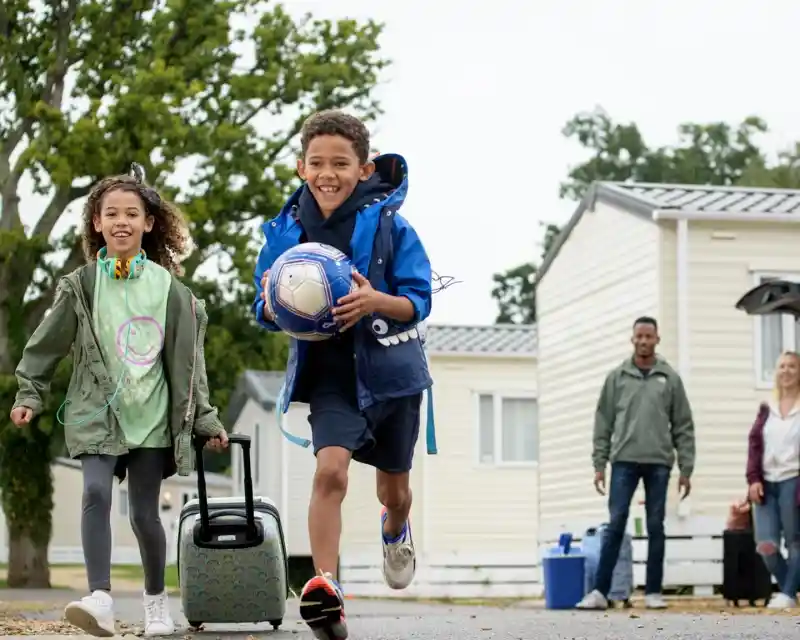 Two children are joyfully running down a path; a boy carries a soccer ball while wearing a blue jacket and shorts. A girl follows, pulling a suitcase and wearing a green shirt with a smiley face. In the background, two adults stand by a parked car near mobile homes.