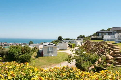 A scenic view of a coastal area featuring several white holiday homes surrounded by lush greenery and vibrant yellow flowers. The ocean is visible in the background under a clear blue sky.