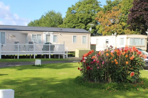 A row of static caravans is set in a lush green area, with a circular flower bed filled with vibrant blooms in the foreground. Tall trees and a clear blue sky complete the tranquil scene.