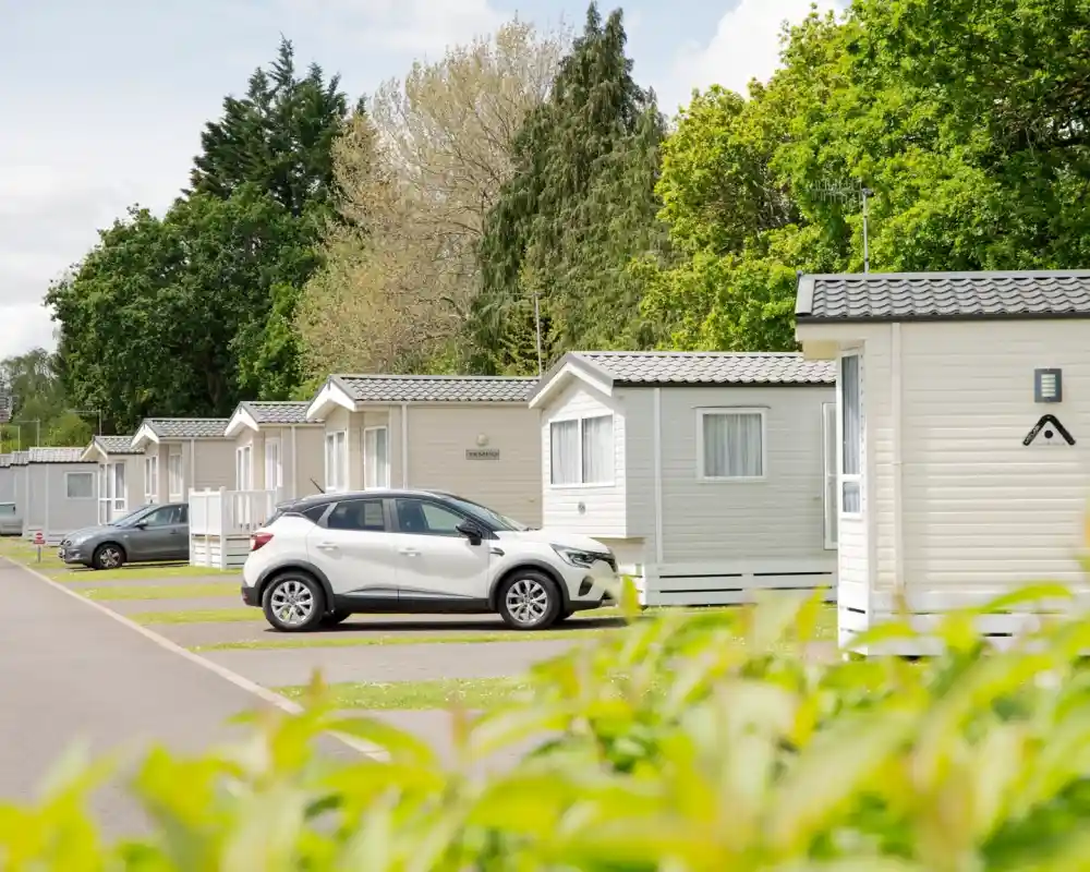 A row of white holiday cabins lined along a neatly paved pathway, with a parked silver car nearby. Lush green trees and bushes frame the scene, indicating a pleasant outdoor setting.