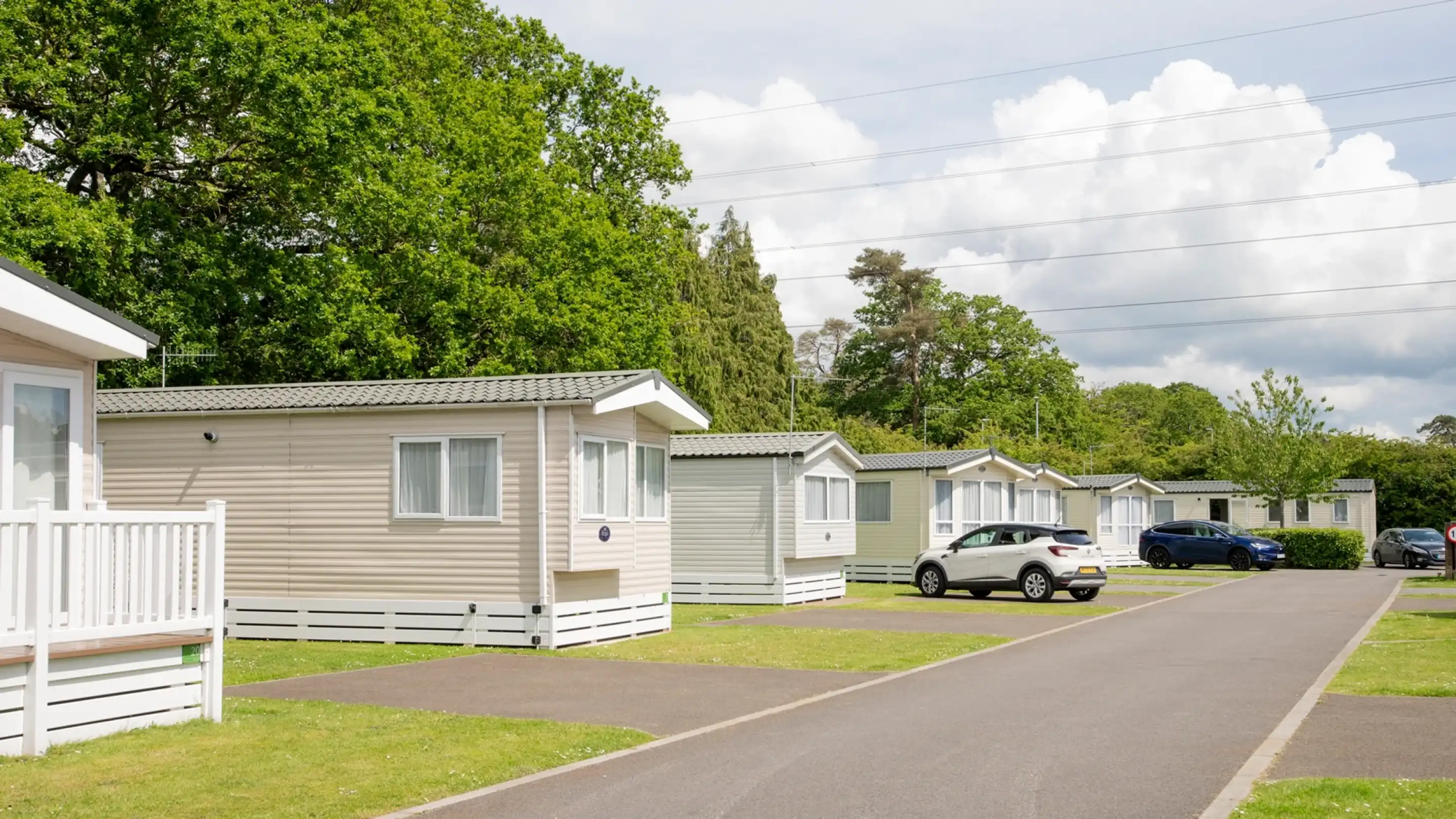 A row of static holiday homes with green lawns alongside a paved road. Trees and a cloudy sky are visible in the background.