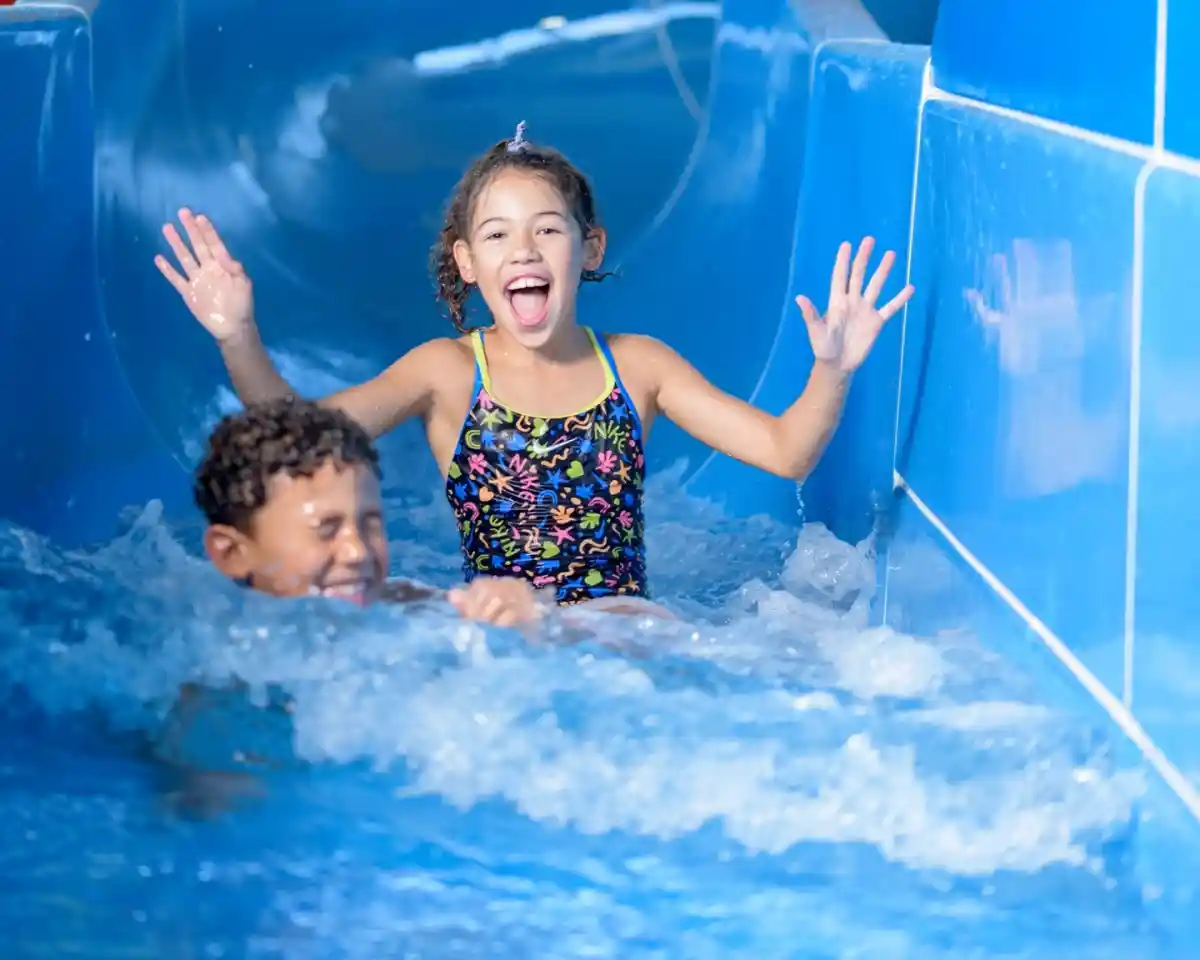 A girl in a colorful swimsuit joyfully slides down a water slide, splashing water around her. A boy swims happily behind her, both enjoying a day at the water park.