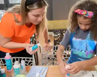 A woman in a bright orange shirt assists a young girl with curly hair, wearing a colorful headband, as they engage in a creative project involving paint and supplies at a table.