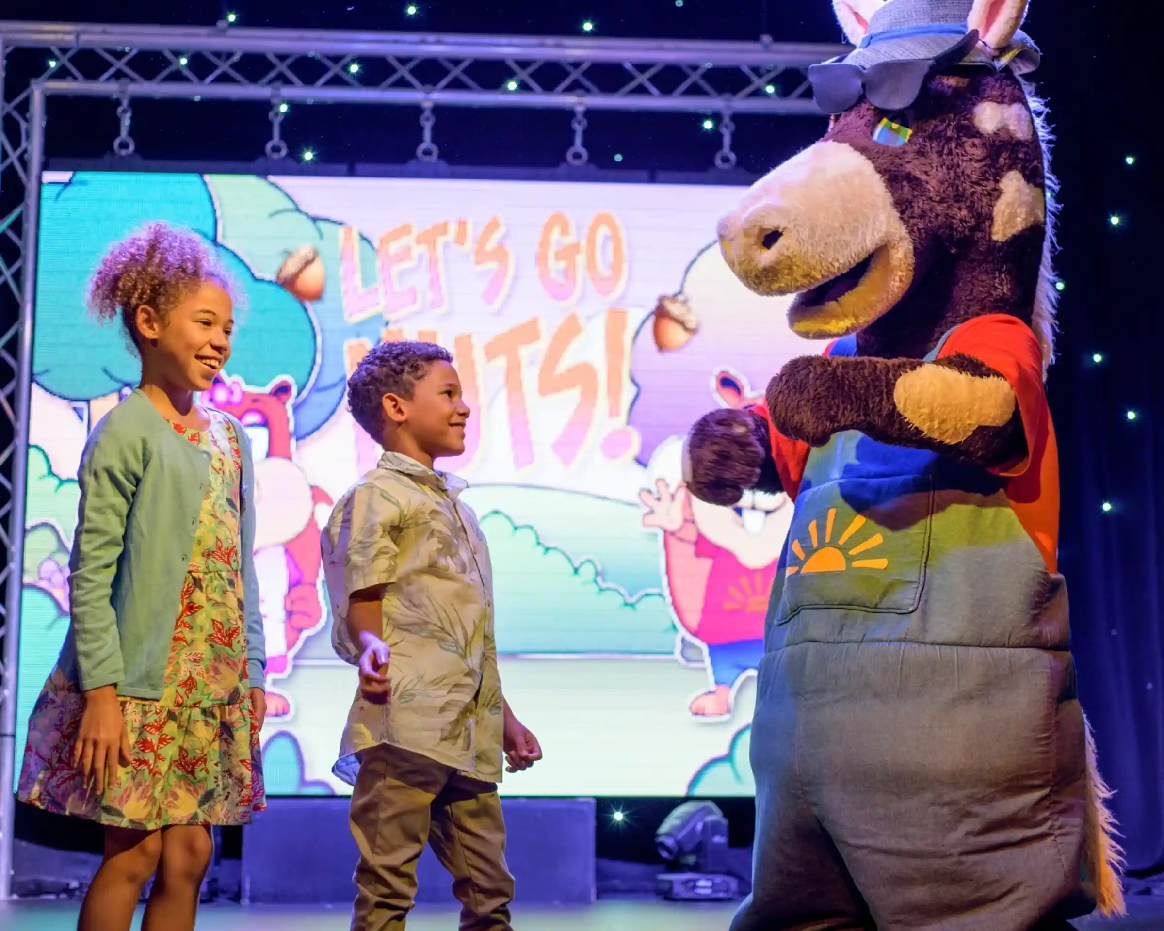 A cheerful mascot dressed as a donkey entertains two smiling children on stage. The backdrop features colorful animated graphics with the text "Let's Go Nuts!"