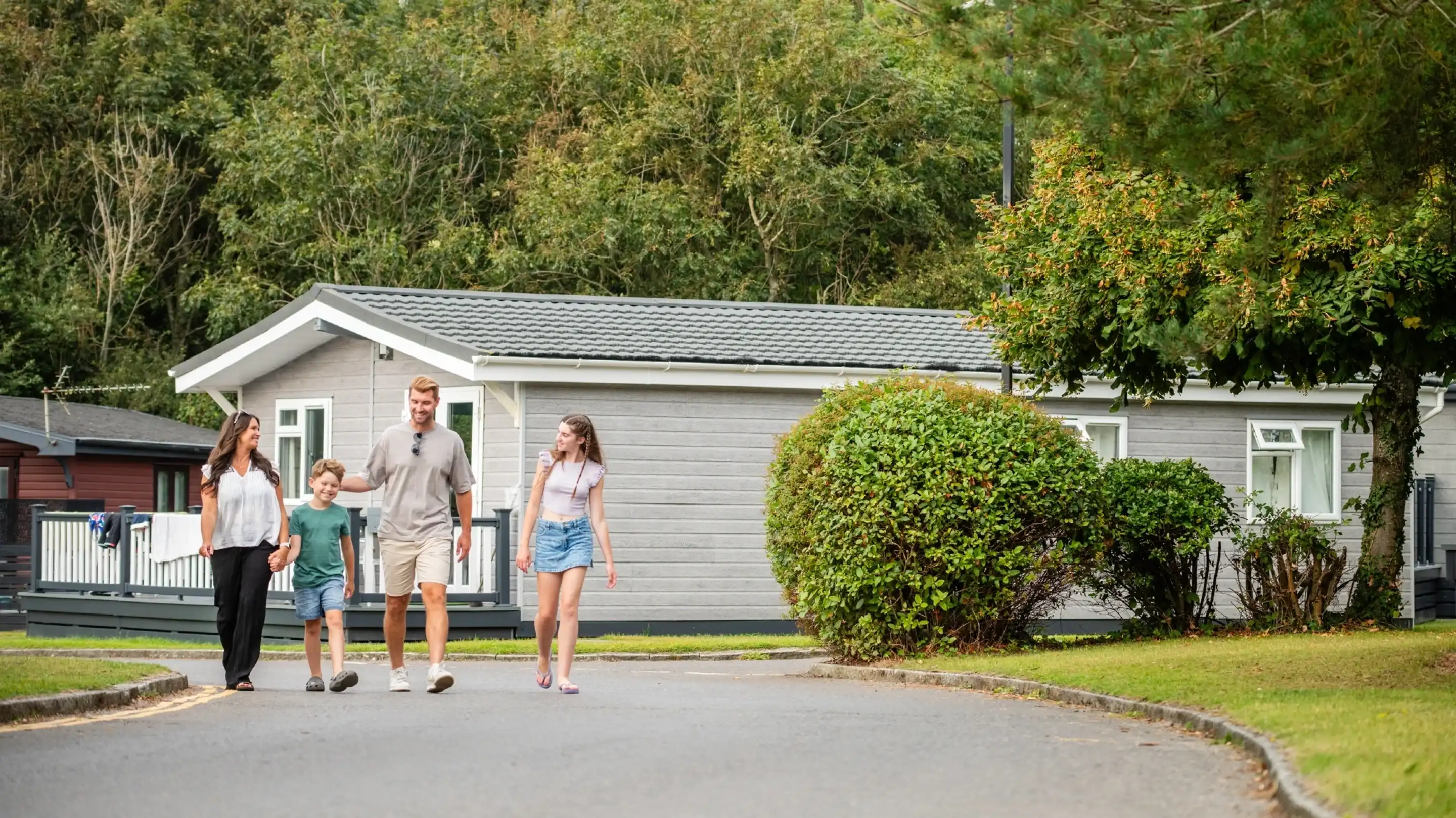 A family of four walks together along a peaceful path, with a modern gray house in the background. The parents, a woman and a man, are accompanied by two children, a boy and a girl, who appear happy as they enjoy their time outdoors. Lush greenery surrounds the area, enhancing the tranquil atmosphere.