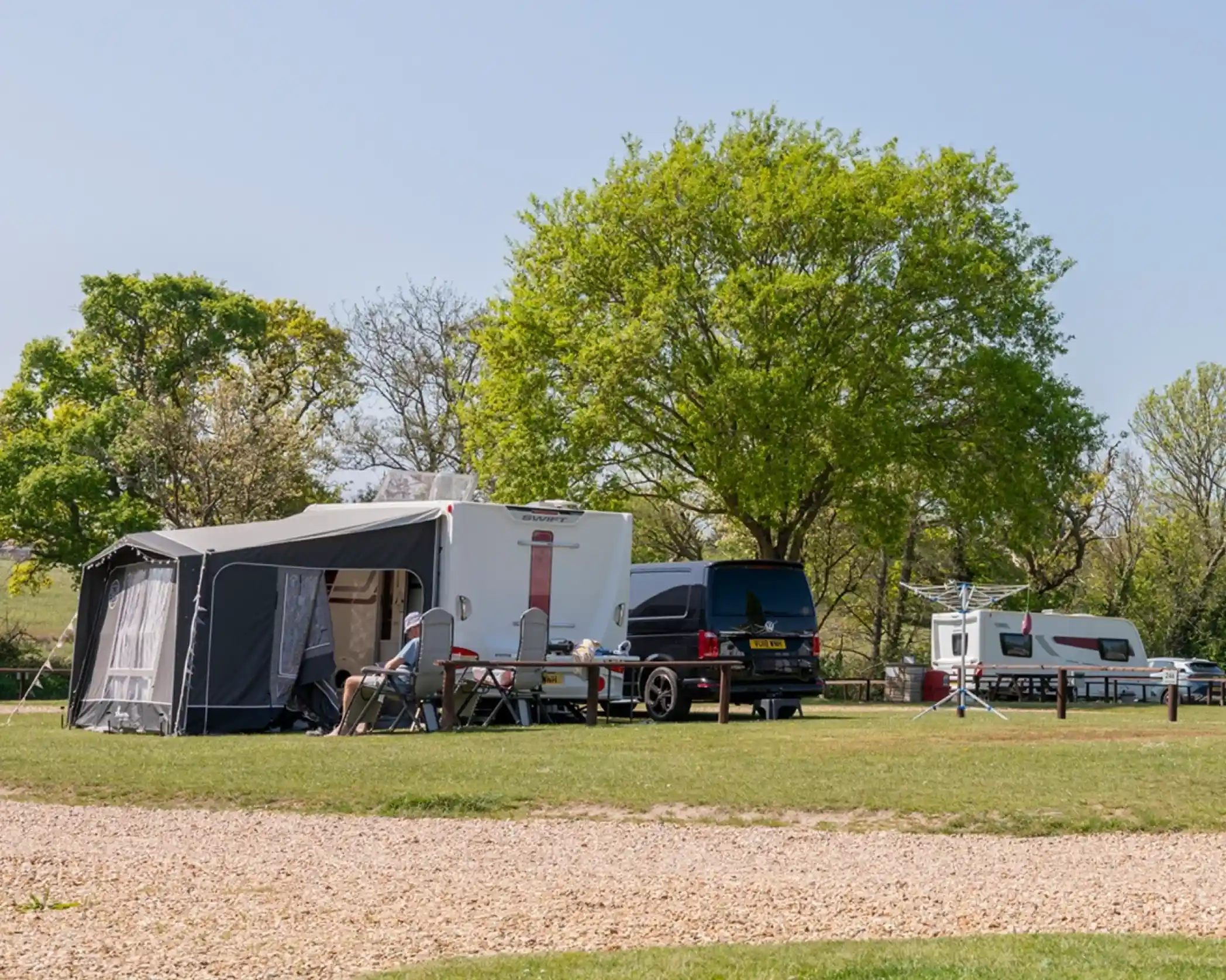 A campsite scene featuring a large trailer with an attached awning, a black van nearby, and a smaller trailer in the background. Green trees and grass create a scenic outdoor setting. A person is seated at a table outside, enjoying the surroundings.