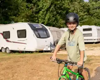 A young boy wearing a helmet smiles while riding a green bicycle near a camping area with caravans in the background. Sunlight illuminates the scene, highlighting the boy's joy and the outdoor setting.