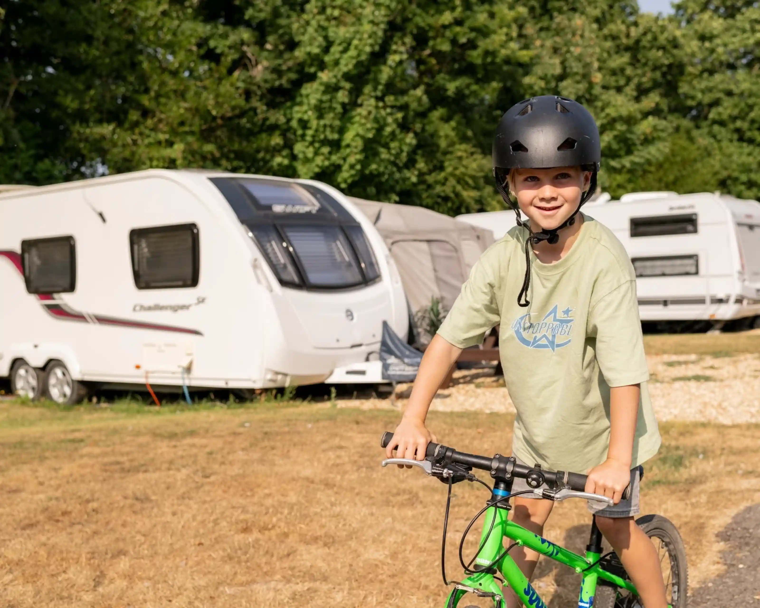 A young boy wearing a helmet smiles while riding a green bicycle near a camping area with caravans in the background. Sunlight illuminates the scene, highlighting the boy's joy and the outdoor setting.