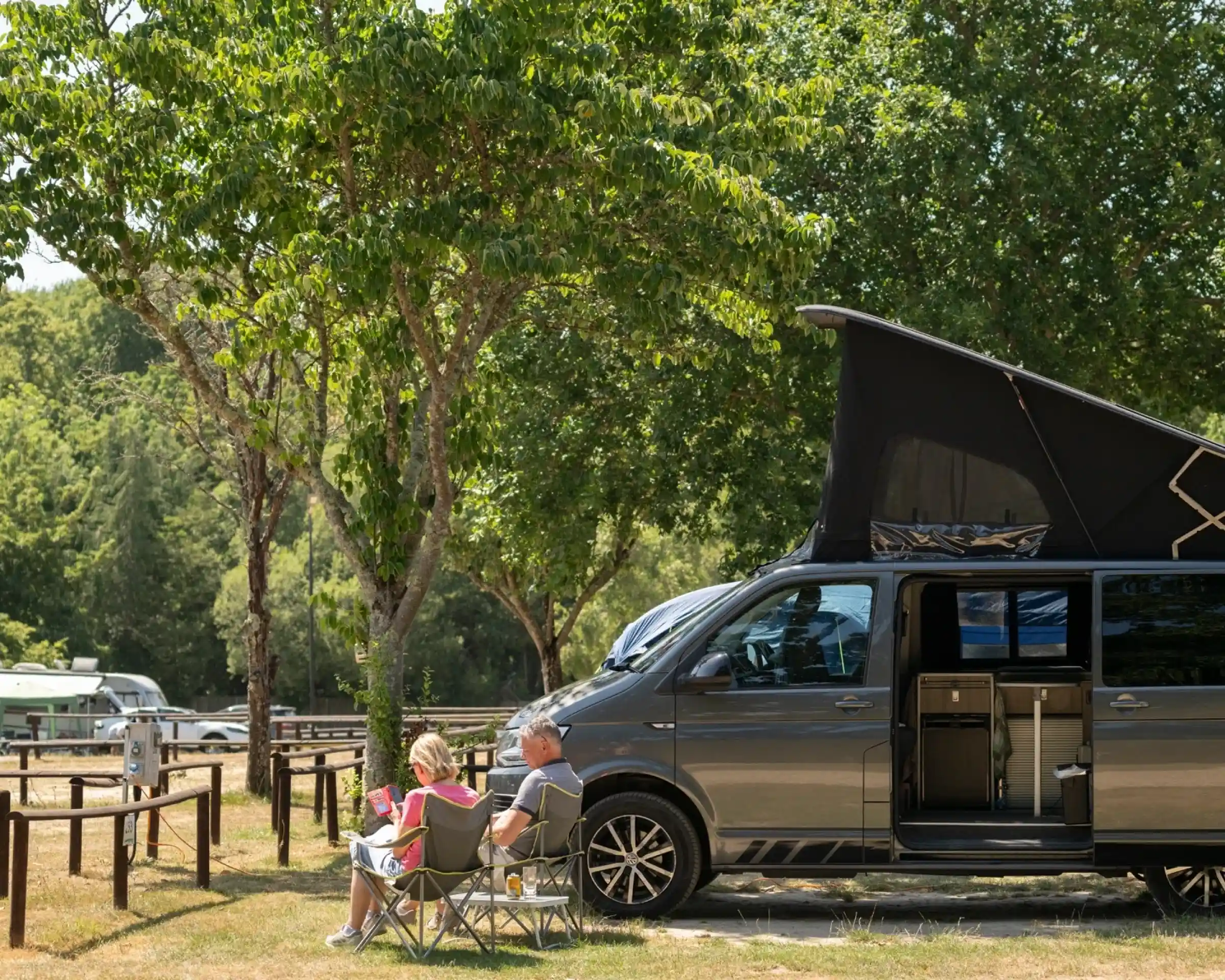 A couple relaxes in camping chairs beside a gray camper van, which has its top raised. They are in a green park area with trees and other campers in the background, enjoying a sunny day.