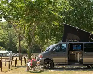 A couple relaxes in camping chairs beside a gray camper van, which has its top raised. They are in a green park area with trees and other campers in the background, enjoying a sunny day.