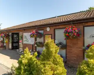 A brick building with large windows adorned with colorful flower baskets. A sign displays information about the location. Bright green shrubs are in the foreground, and the sky is clear blue.