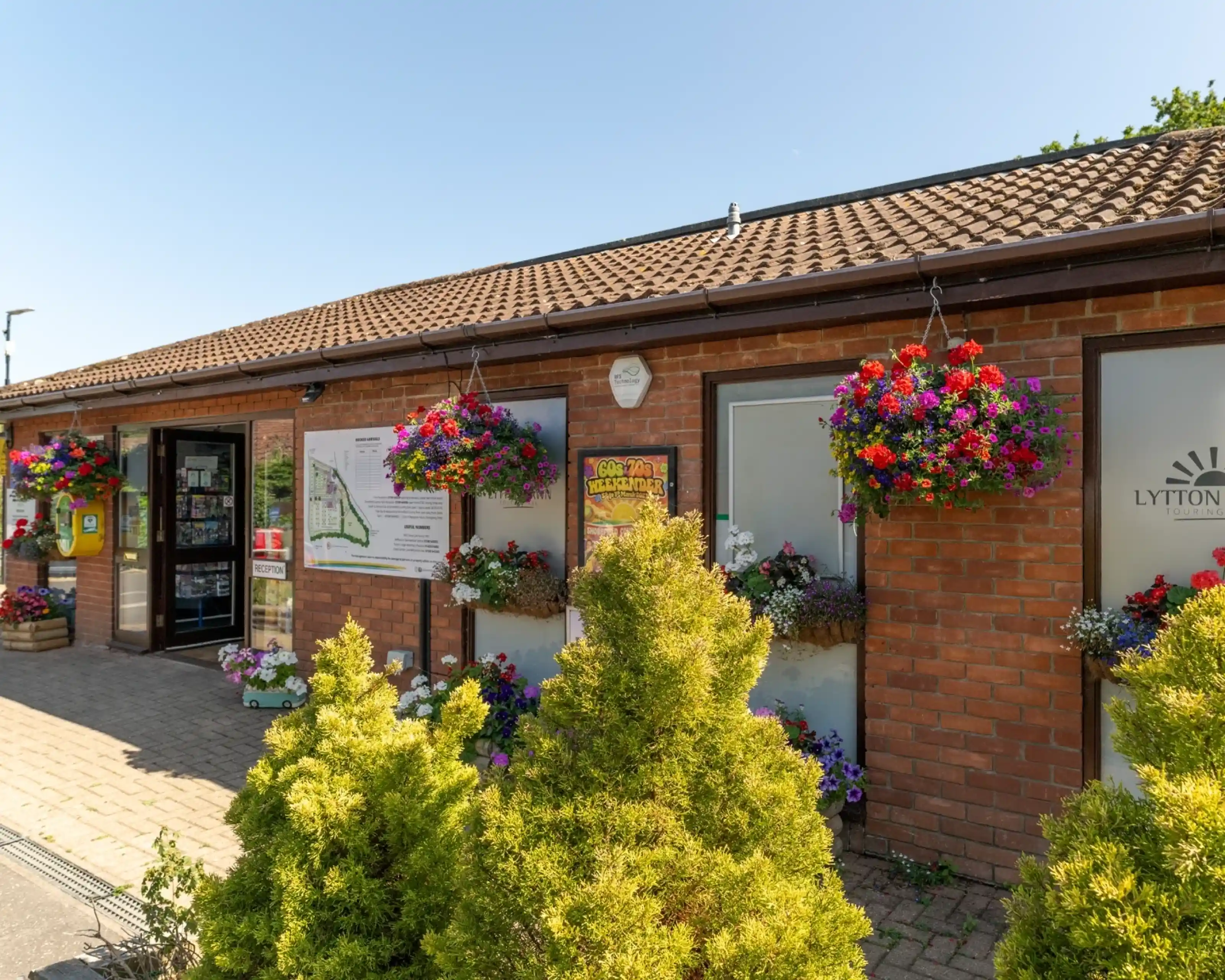 A brick building with large windows adorned with colorful flower baskets. A sign displays information about the location. Bright green shrubs are in the foreground, and the sky is clear blue.