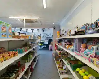 A brightly lit aisle in a shop, lined with shelves filled with various groceries and toys. A checkout area is visible in the background, adorned with banners. Items include snacks, canned goods, and children's games.