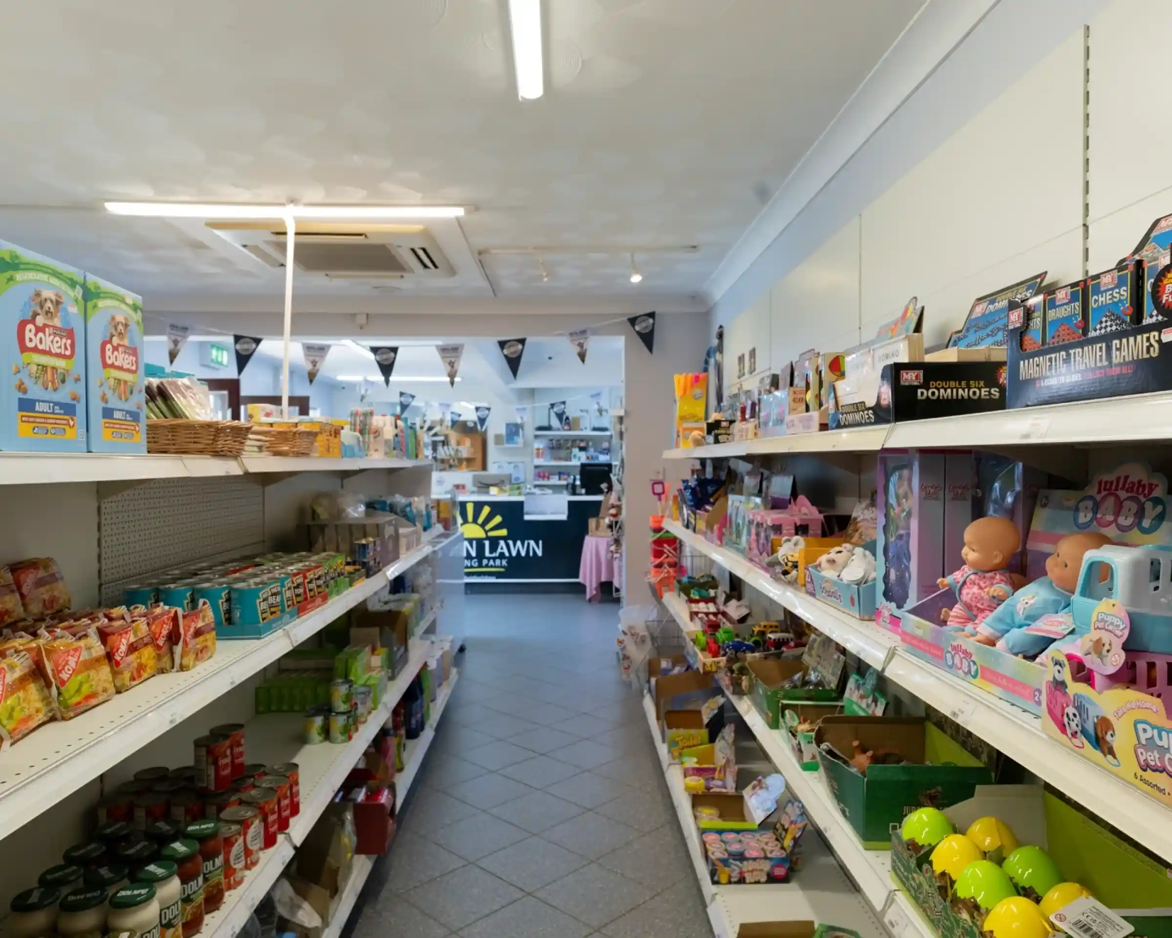 A brightly lit aisle in a shop, lined with shelves filled with various groceries and toys. A checkout area is visible in the background, adorned with banners. Items include snacks, canned goods, and children's games.