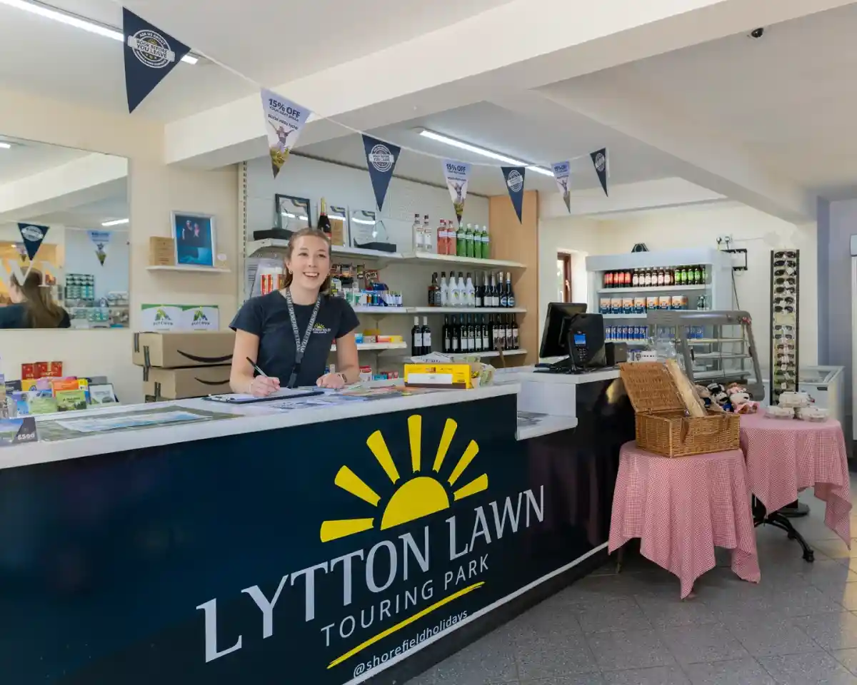 A smiling staff member stands behind the reception counter at Lytton Lawn Touring Park, surrounded by shelves filled with various products. The area features a bright and welcoming atmosphere, with bunting hanging from the ceiling and a display of drinks and snacks nearby.