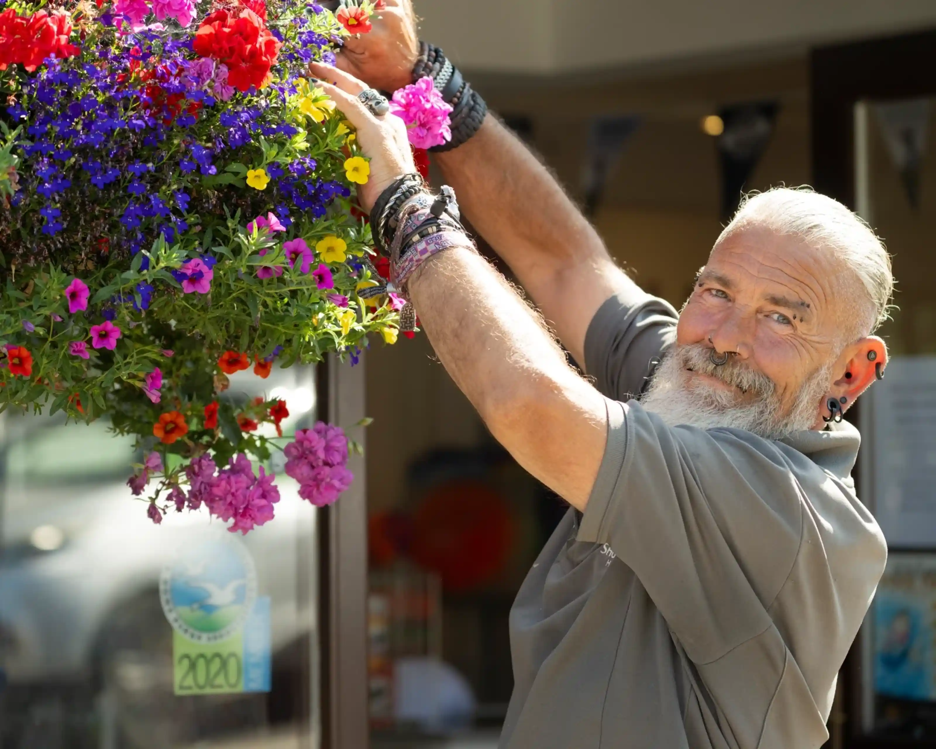 A smiling elderly man with a gray beard and earrings hangs a vibrant flower basket filled with colorful petunias and other blooms outside a shop.