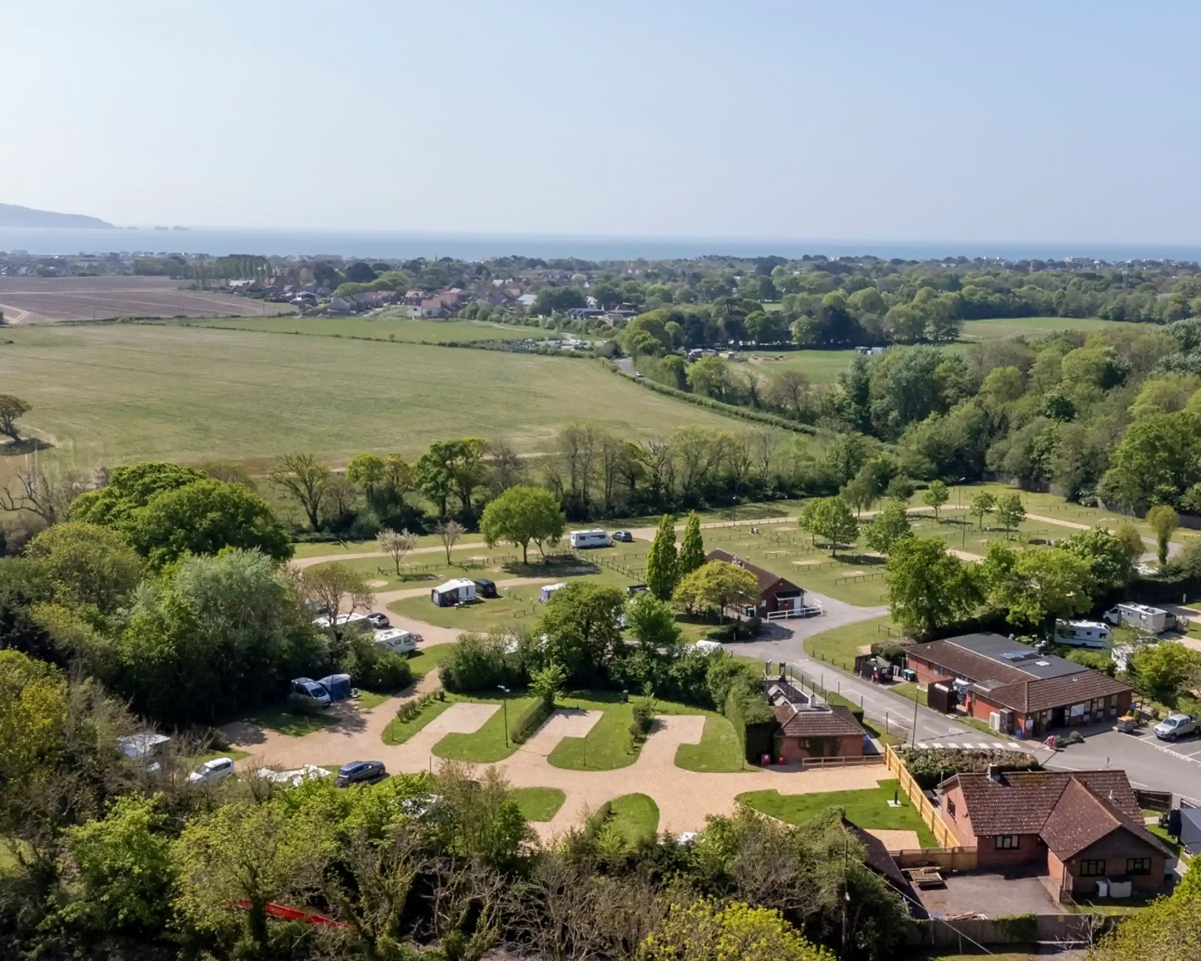 Aerial view of a scenic campsite with lush greenery, parked caravans, and trees. In the background, rolling hills and a distant coastline lead to the sea under a clear blue sky.