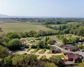 Aerial view of a scenic campsite with lush greenery, parked caravans, and trees. In the background, rolling hills and a distant coastline lead to the sea under a clear blue sky.