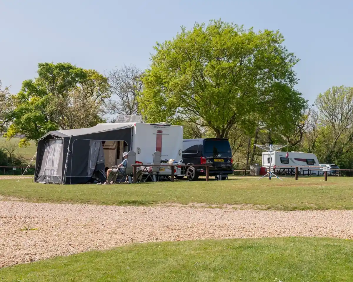 A campsite scene featuring a large camper with an awning, a parked black van, and a caravan, surrounded by lush green trees and a grassy area. Wooden picnic tables are nearby.