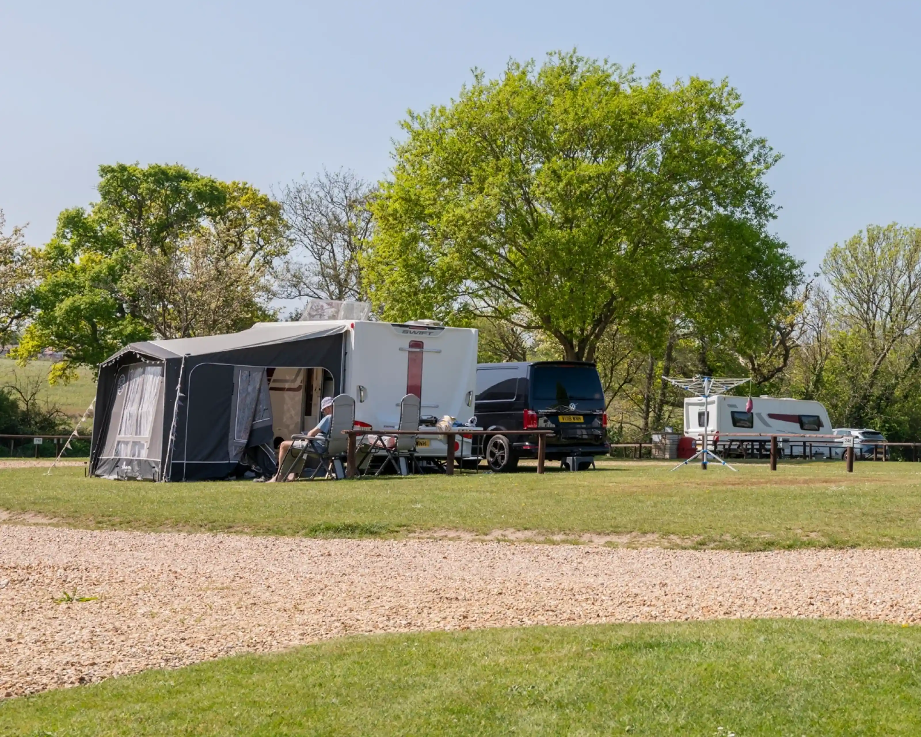 A campsite scene featuring a large camper with an awning, a parked black van, and a caravan, surrounded by lush green trees and a grassy area. Wooden picnic tables are nearby.