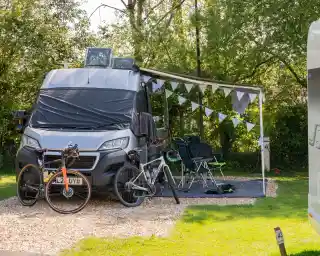 A camper van is parked on a gravel site, with an awning extended and outdoor chairs set up. Two bicycles are leaning against the van, and decorative bunting hangs nearby among lush green trees.