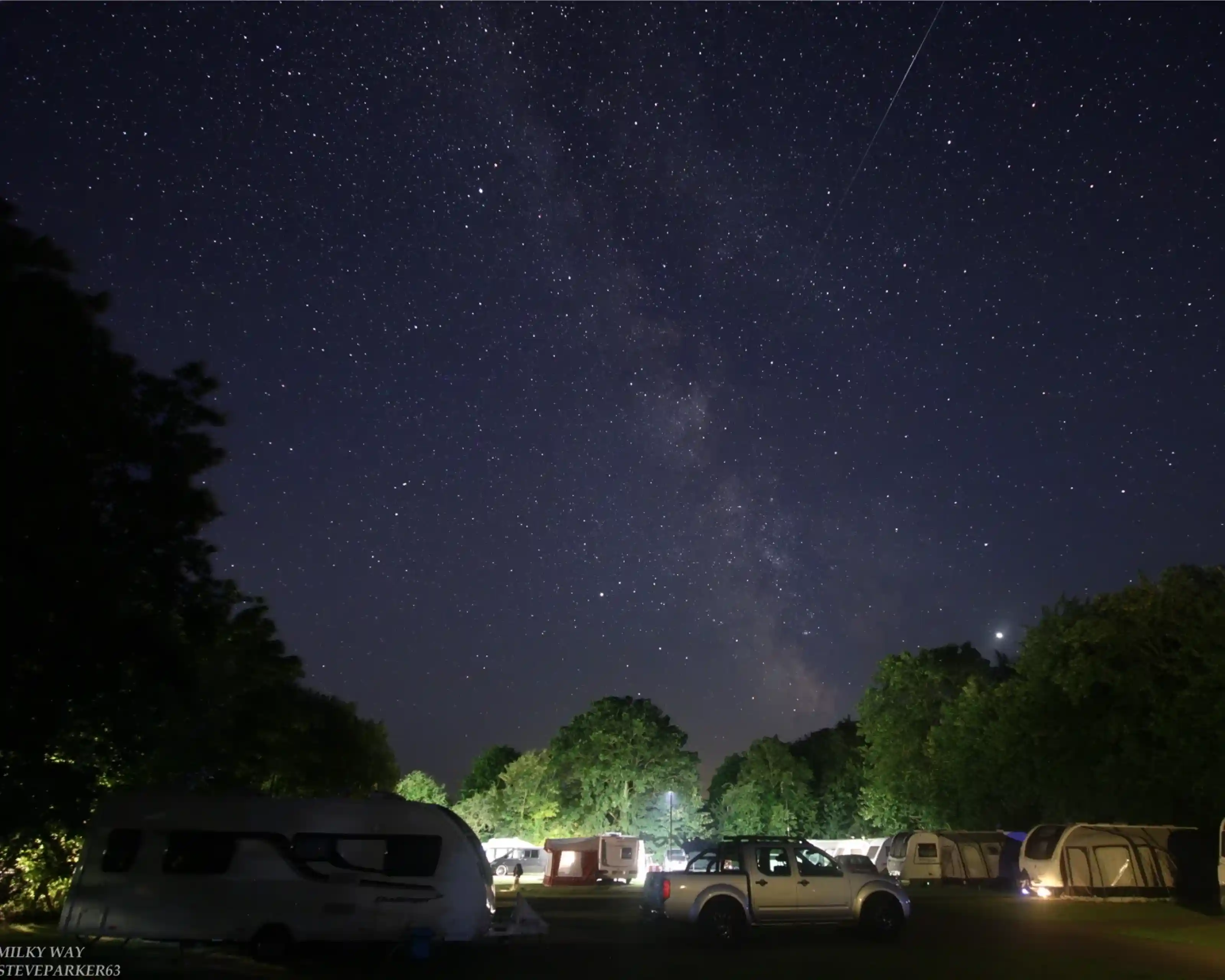 A starry night sky features the Milky Way above a campsite with parked caravans and trees. Subtle lights from the caravans illuminate the scene, creating a serene atmosphere.