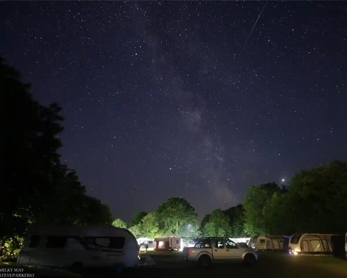 A starry night sky features the Milky Way above a campsite with parked caravans and trees. Subtle lights from the caravans illuminate the scene, creating a serene atmosphere.