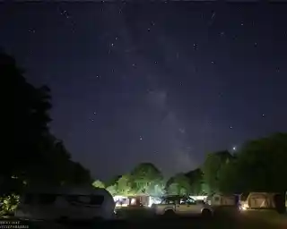 A starry night sky features the Milky Way above a campsite with parked caravans and trees. Subtle lights from the caravans illuminate the scene, creating a serene atmosphere.