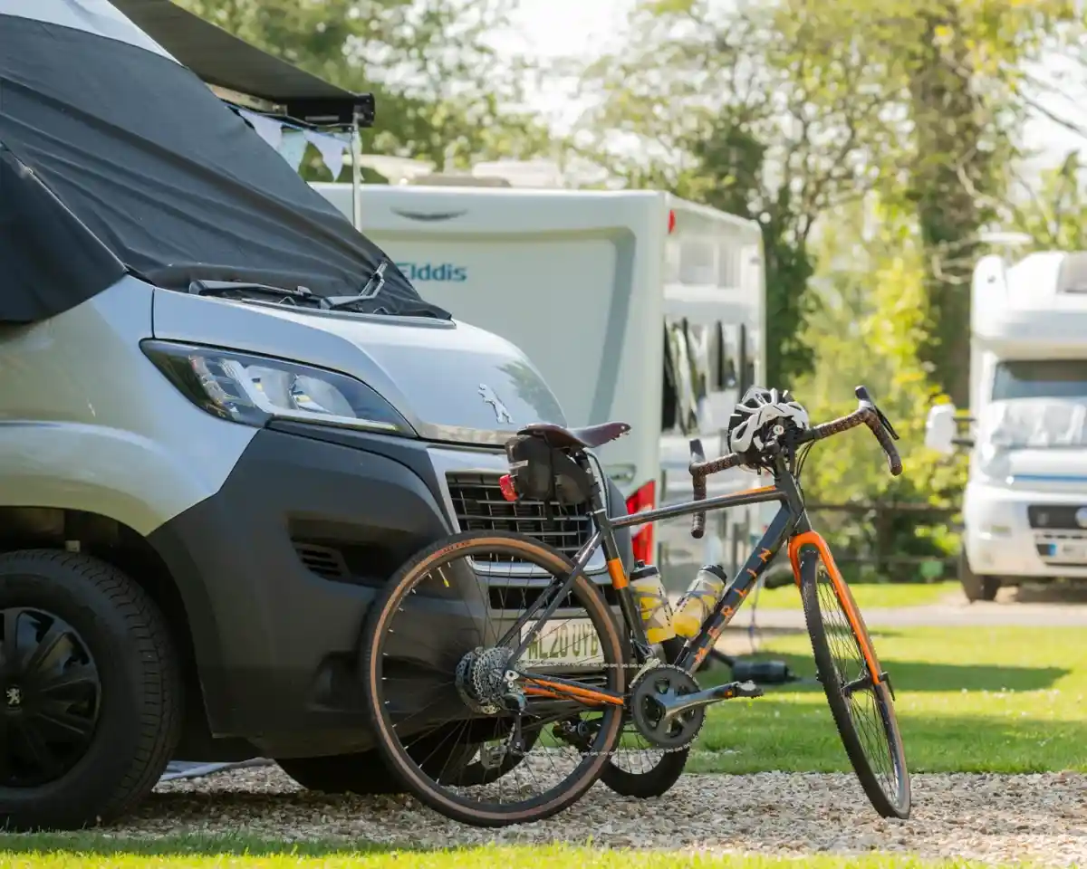 A bicycle leans against a camper van in a grassy area. Several other camper vans are parked in the background, surrounded by trees and greenery.