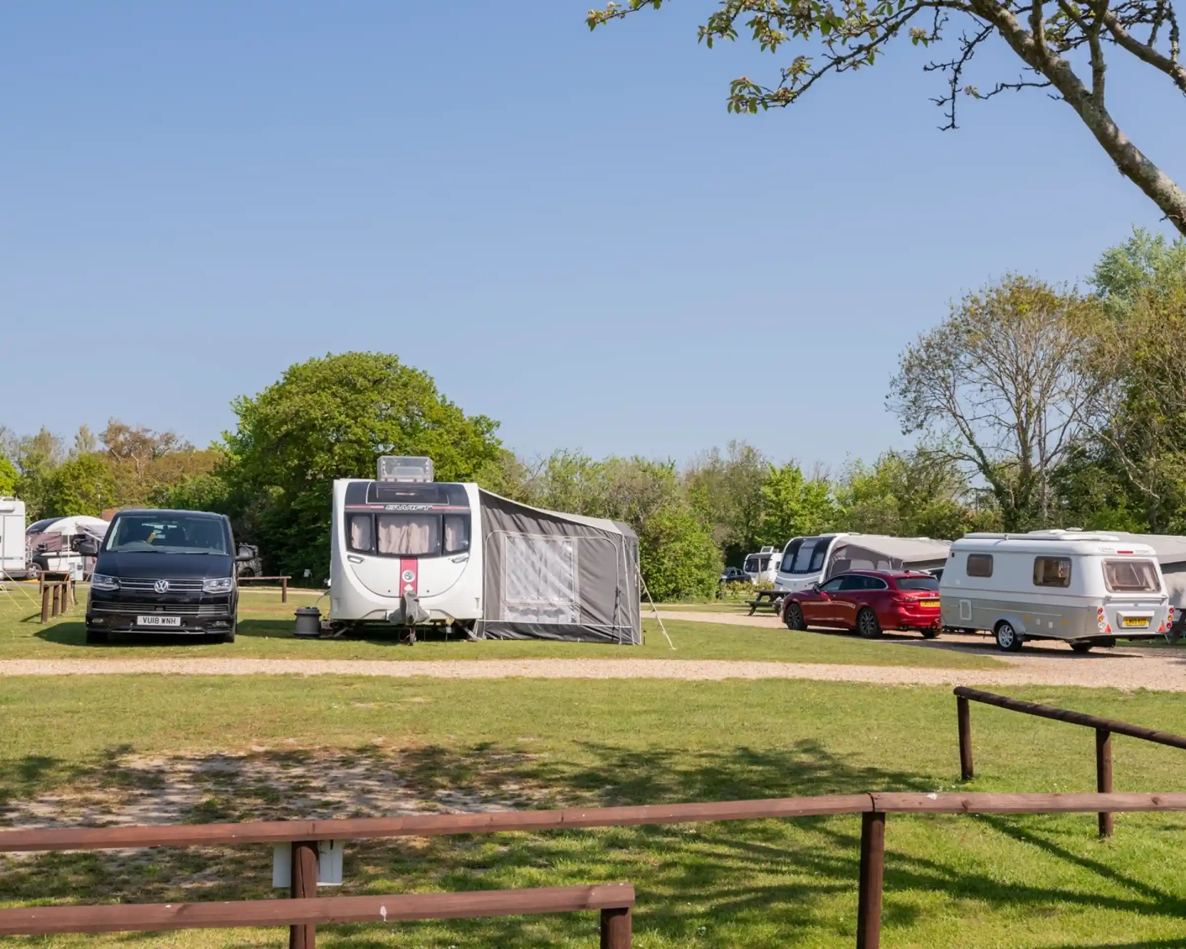 A campsite featuring several caravans and camper vans set against a clear blue sky. Lush greenery surrounds the area, while a wooden fence runs in the foreground.