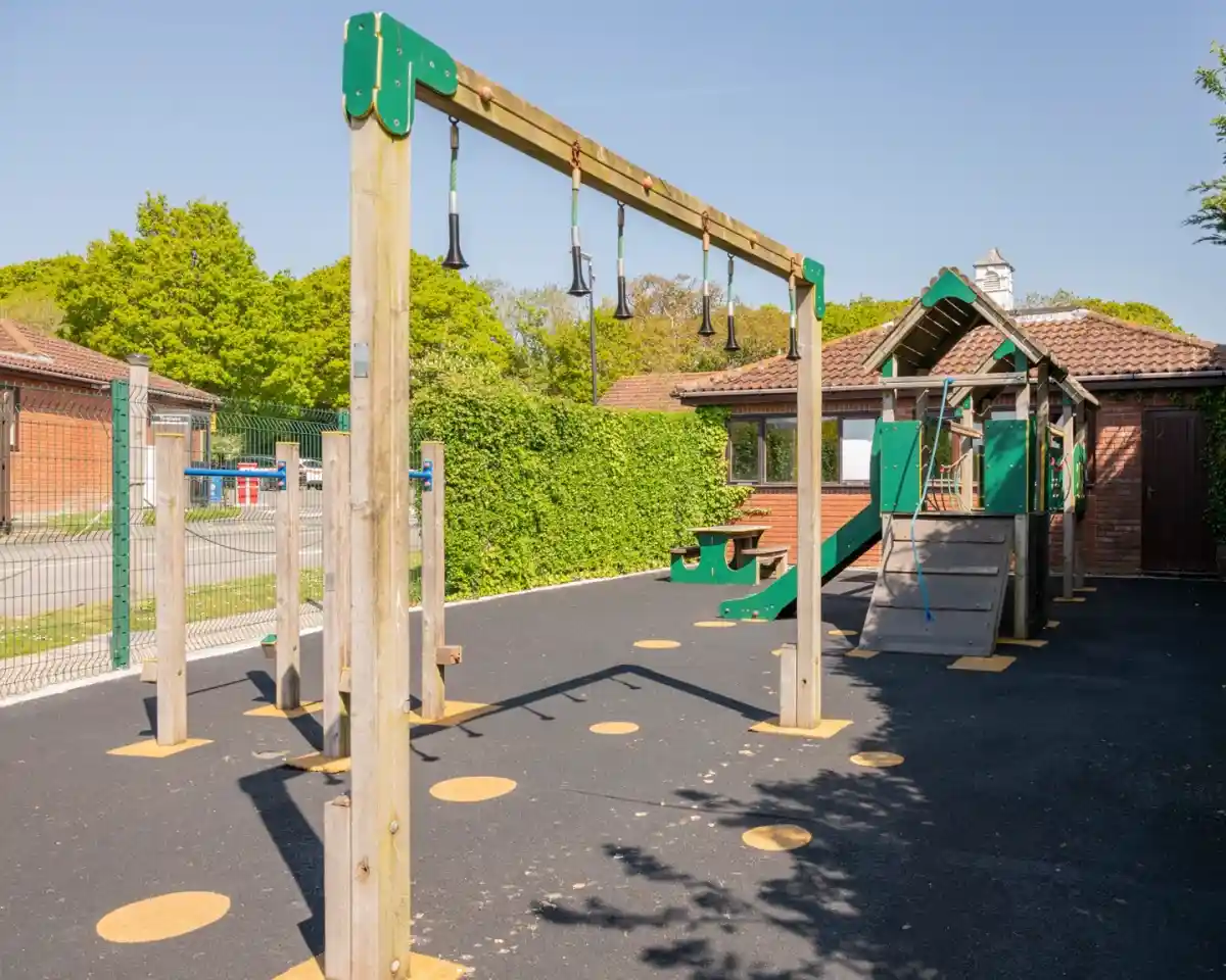 A playground with swings and a climbing structure, surrounded by green trees and a blue sky. The area features a black rubber surface and wooden supports for the equipment. A building is visible in the background.